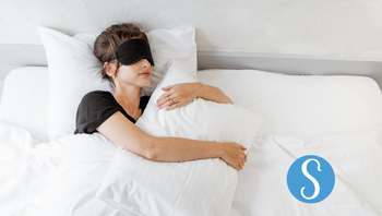 A woman sleeping in bed with an eye mask on. Her mouth is closed and she's holding a pillow.