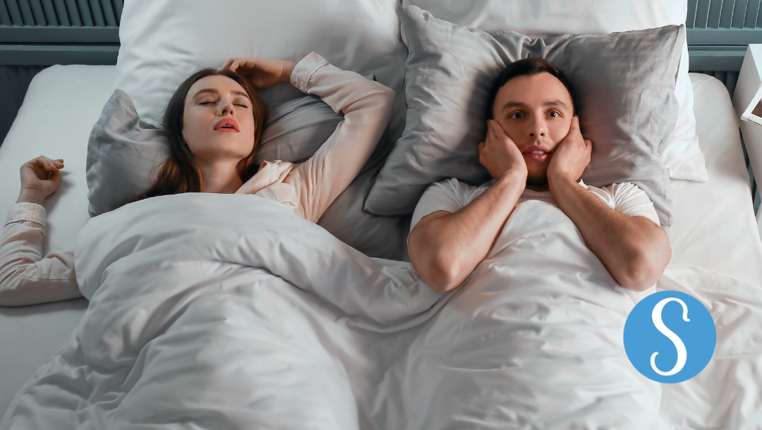 A couple lying in bed. The man is trying to block the sound of the women's snoring.